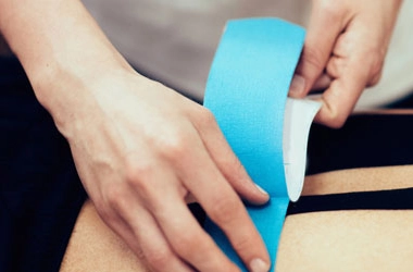 Close-up of hands applying a strip of blue kinesiology tape to a person’s back