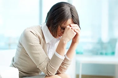 Person sitting indoors on a white chair, leaning forward with one hand resting on the forehead. The individual is wearing a light-colored sweater over a white collared shirt