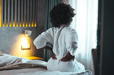 Person wearing a white bathrobe sitting on the edge of a bed, with hands placed on the lower back as if experiencing discomfort