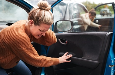Person wearing a brown knitted sweater opening the door of a blue car, with another person in the background near a second car